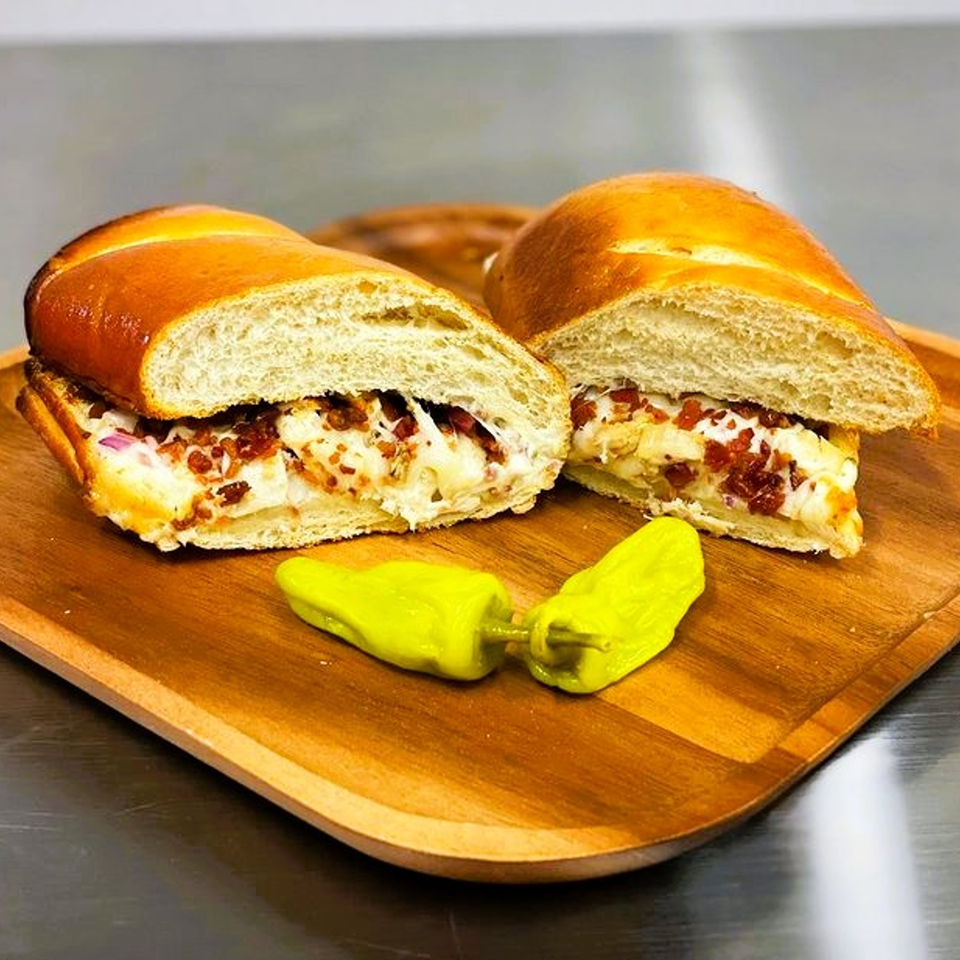 Two halves of a cheesy bacon and onion sub sandwich with two yellow peppers on a wooden board.