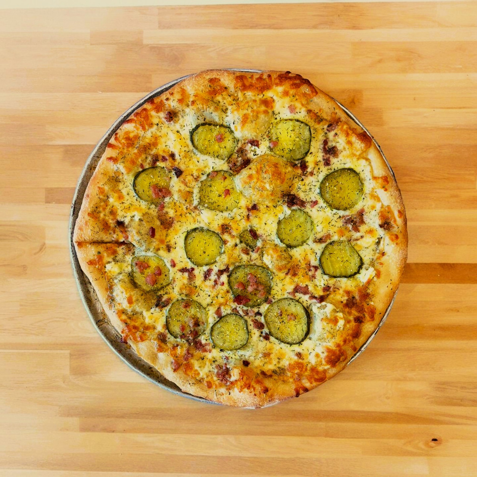 Hatcher Pass Pizza's dill pickle pizza, topped with pickles, bacon, cheese, and dill, on a wooden table.